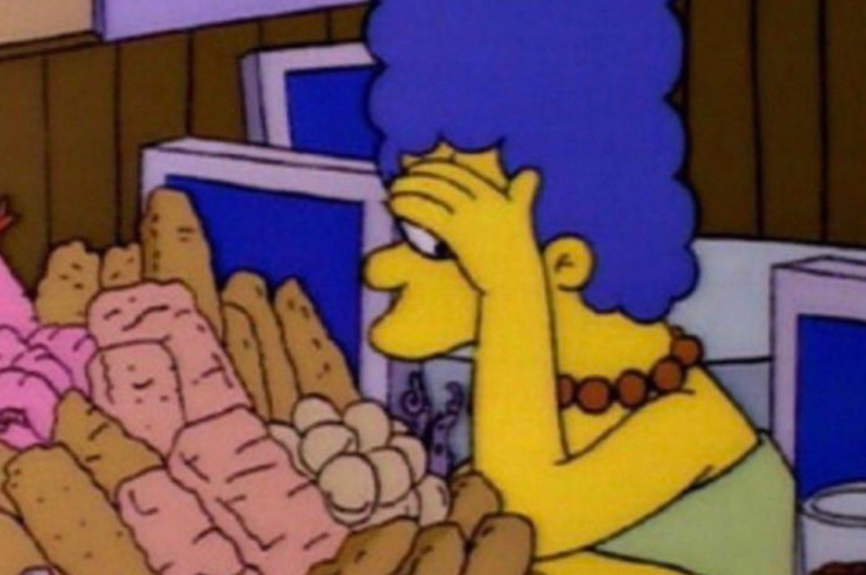Marge Simpson, from The Simpsons, is sitting at a table with her hand over her eyes and a variety of baked goods in front of her
