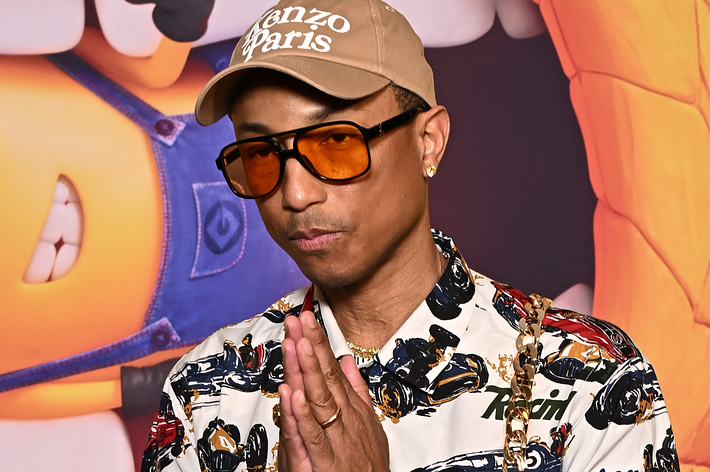 Pharrell Williams wearing a patterned shirt, orange-tinted glasses, and a cap with "Kenzo Paris" written on it, posing with his hands together in front of a colorful background