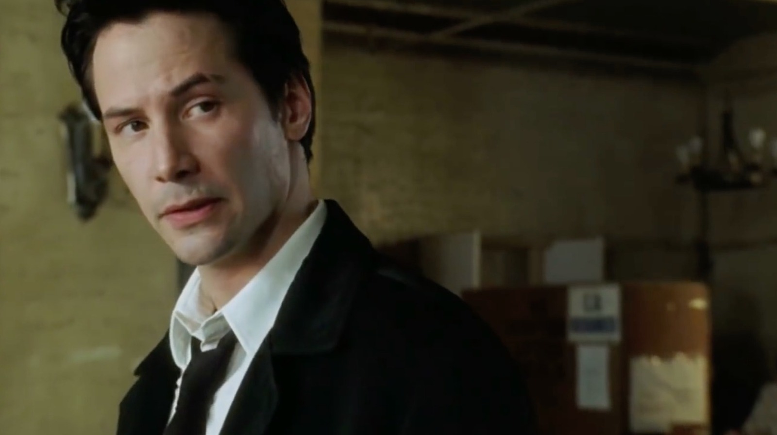 Keanu Reeves, dressed in a suit with a black coat, looks to the side inside a dimly lit room with boxes in the background