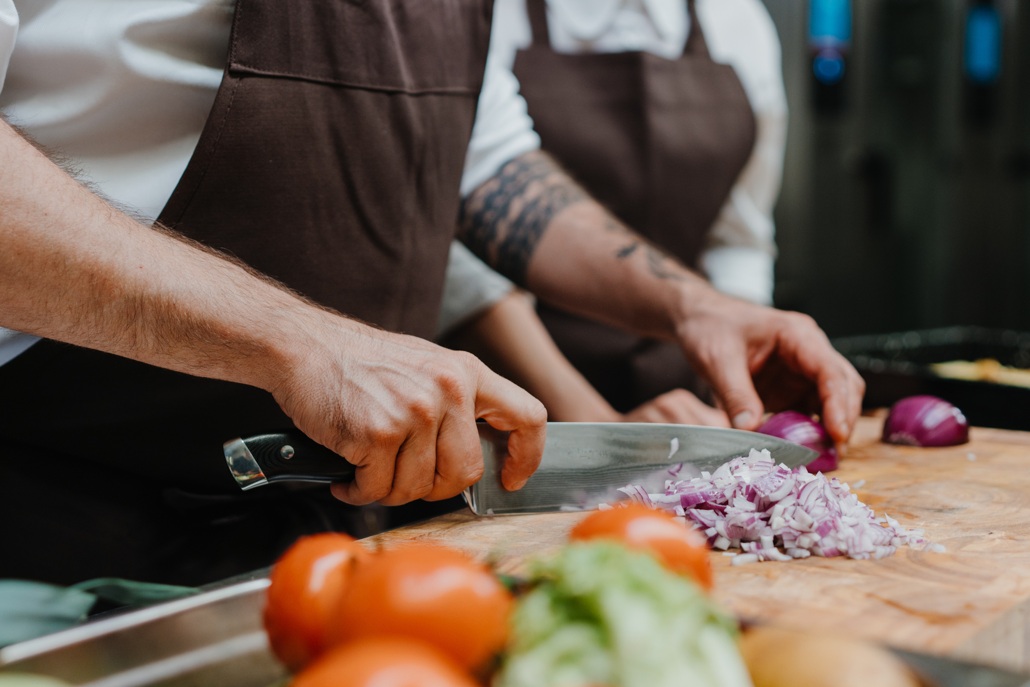 Two chefs in aprons chopping onions and tomatoes on a wooden cutting board in a professional kitchen