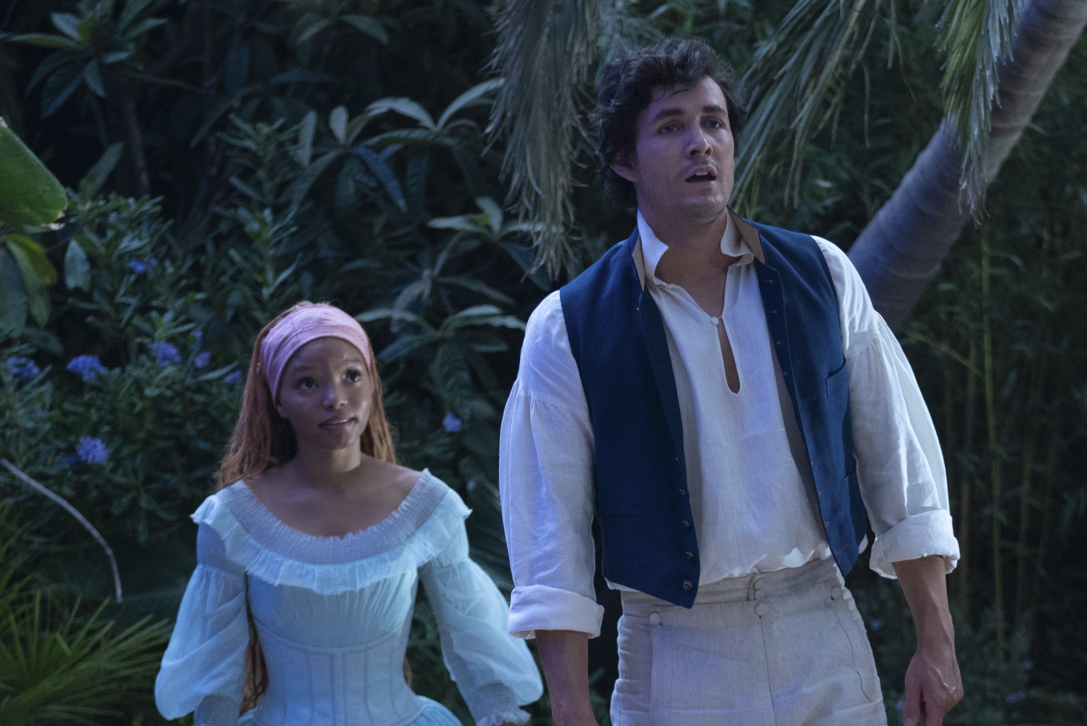 Halle Bailey and Jonah Hauer-King in a wooded setting, both dressed in period costumes; Halle in an off-the-shoulder dress, Jonah in a vest and period shirt