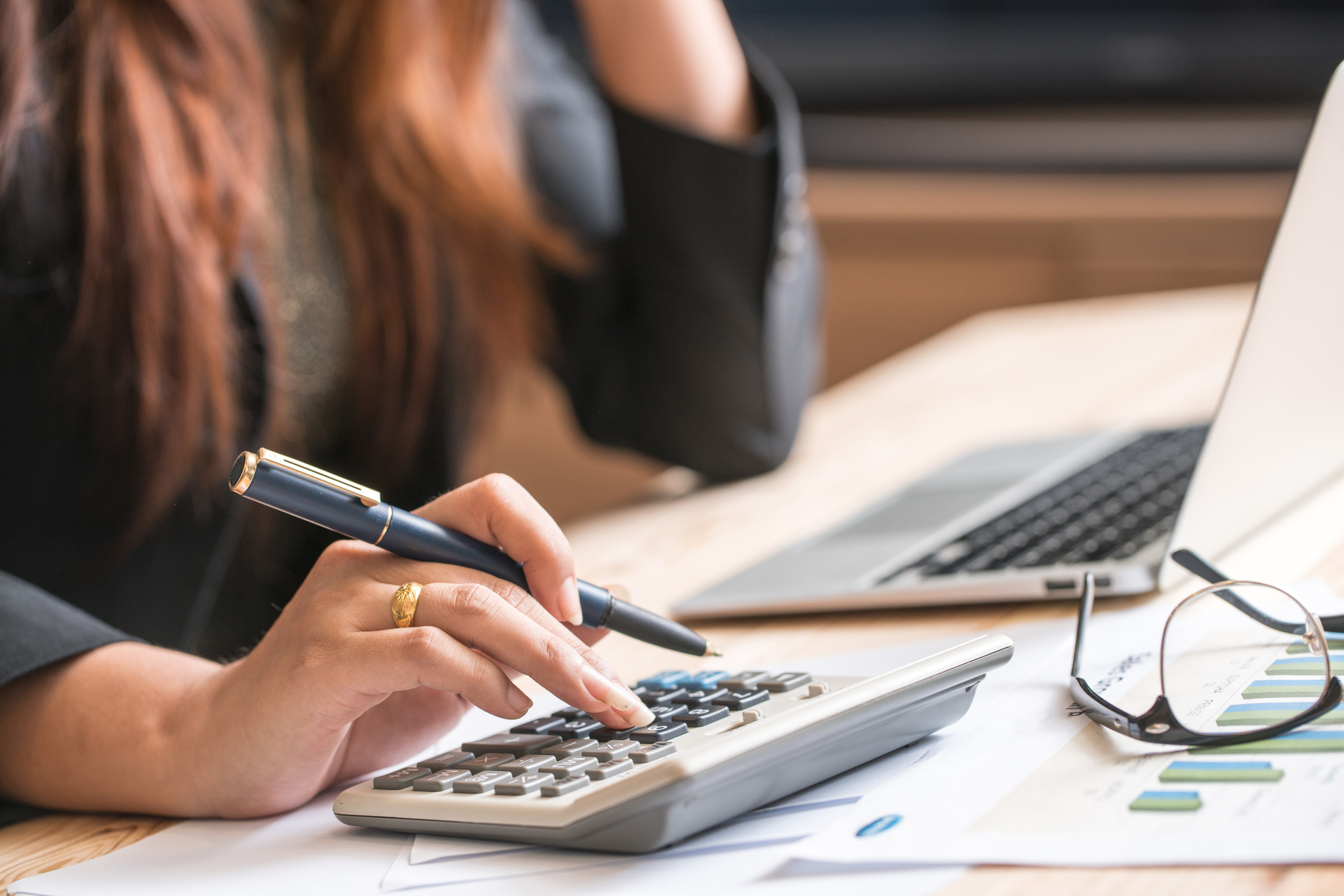 A person using a calculator at a desk with documents and a laptop, possibly engaged in financial or work-related tasks. Glasses and a pen are also visible