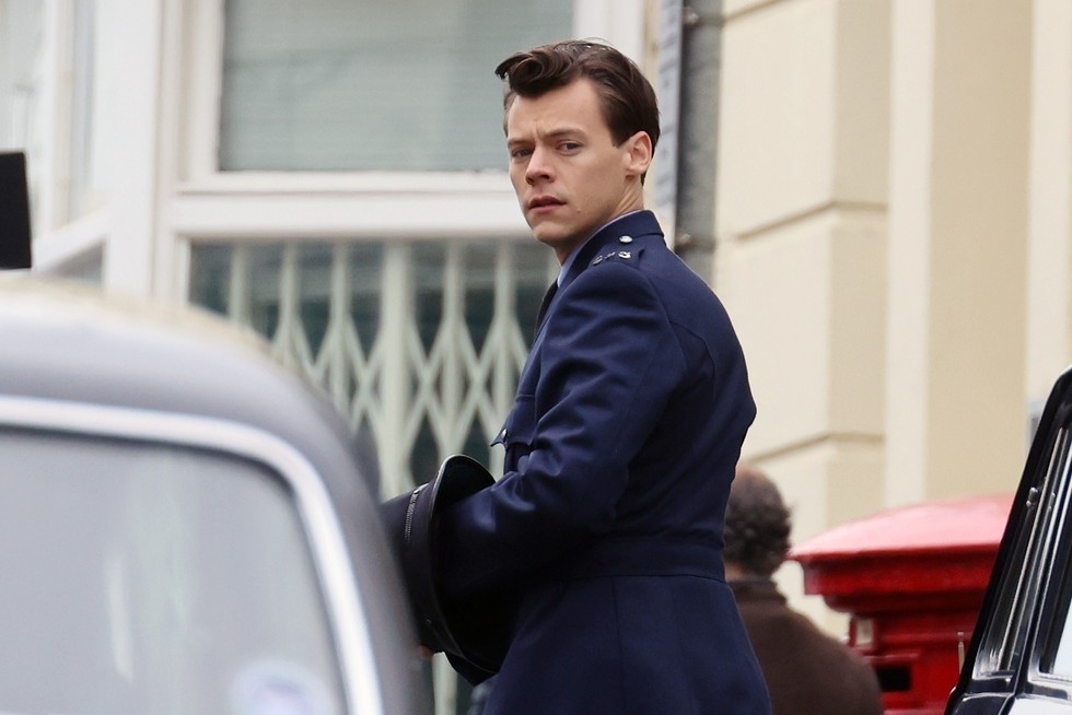 Harry Styles dressed in a blue military-style uniform, holding a hat, looking back while standing near a building with a window and red mailbox