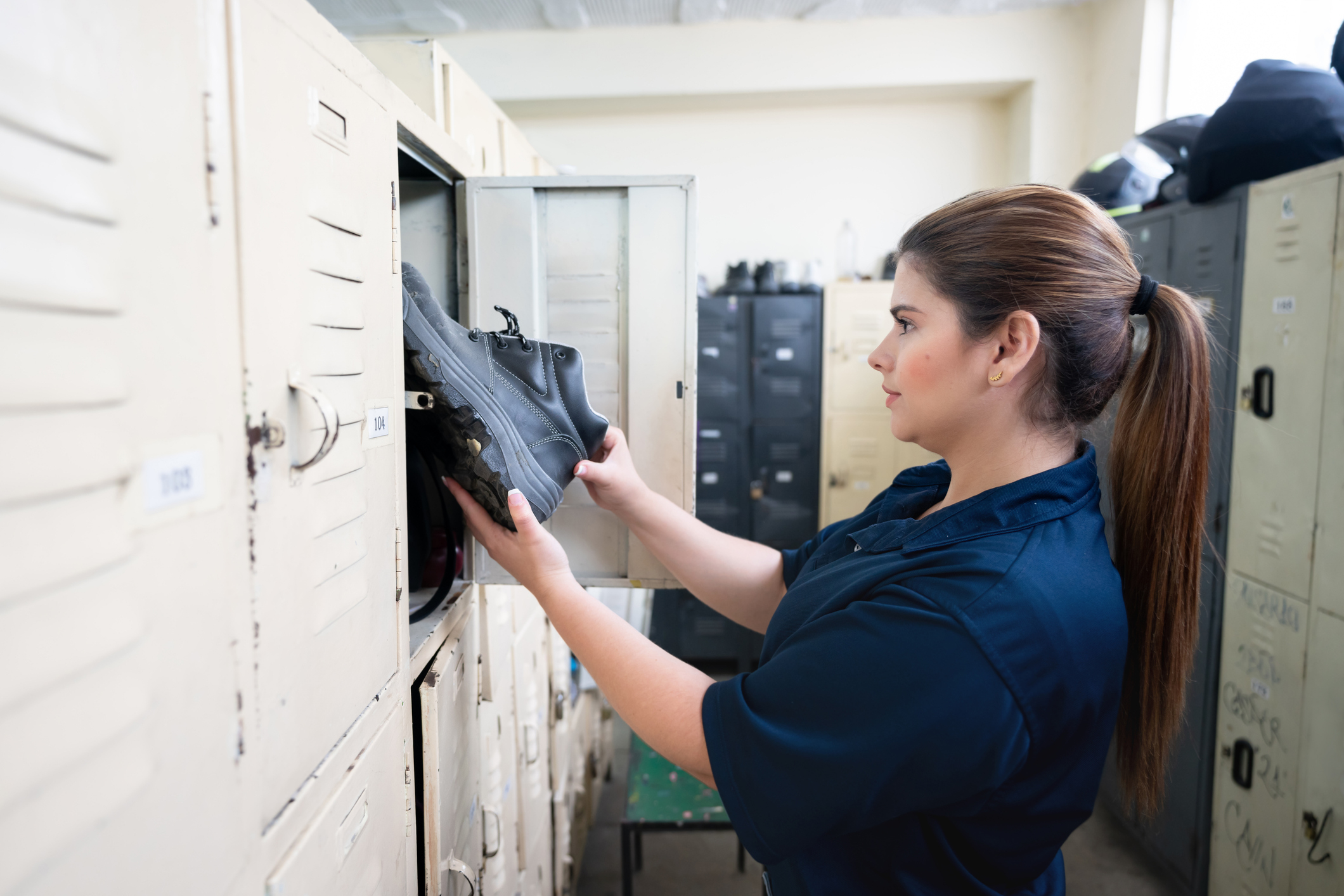 A woman in a work uniform places boots into a locker at her workplace. Lockers are visible in the background