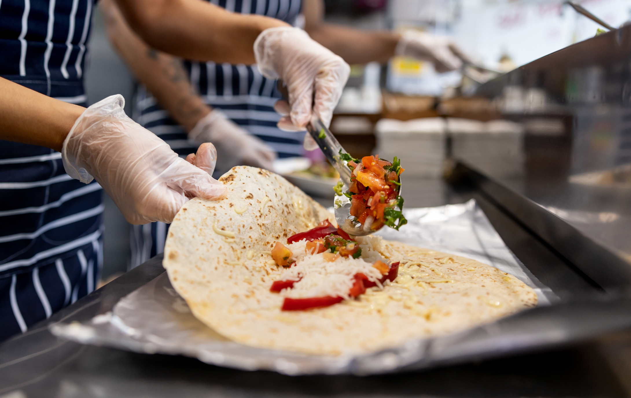Two people wearing striped aprons and gloves are assembling a burrito, adding ingredients like rice, diced tomatoes, and bell peppers at a food service counter