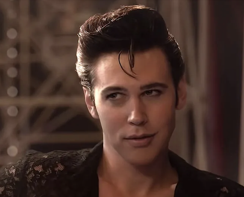 A person with a styled pompadour hairstyle gives a confident look while wearing a lace-patterned top. The background is blurred with lights and structures