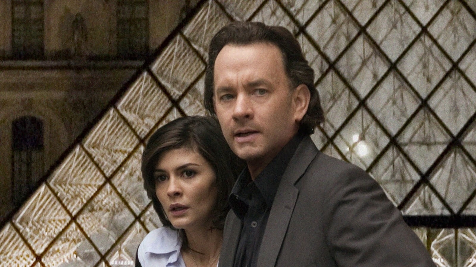 Audrey Tautou and Tom Hanks are standing in front of a large glass pyramid. Both appear focused and attentive