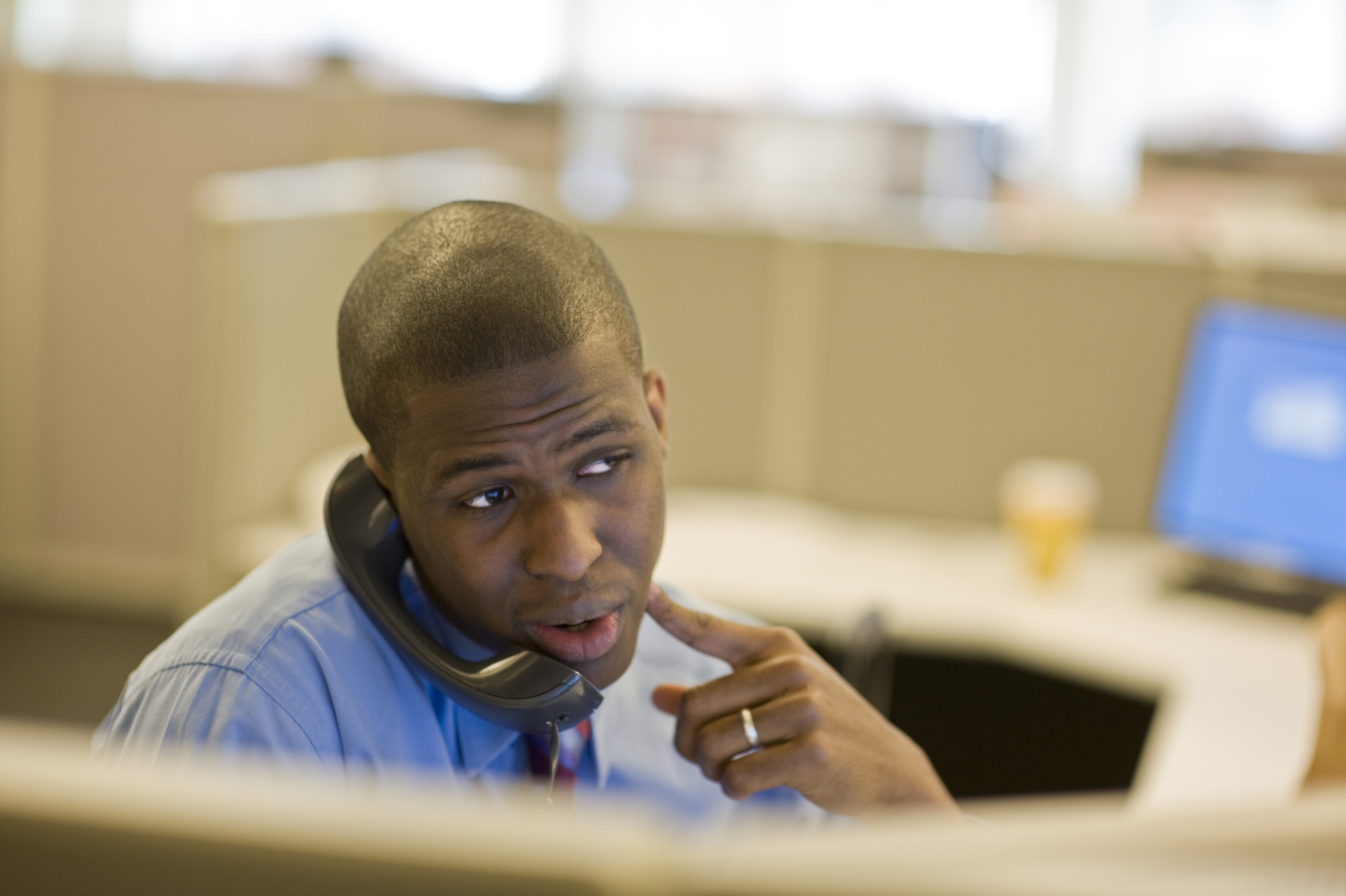 A man in a business setting is speaking on the phone, appearing thoughtful while pointing to his cheek. Office cubicles are visible in the background
