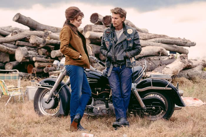 Jodie Comer and Austin Butler dressed in retro outfits, leaning against a vintage motorcycle with stacked logs in the background