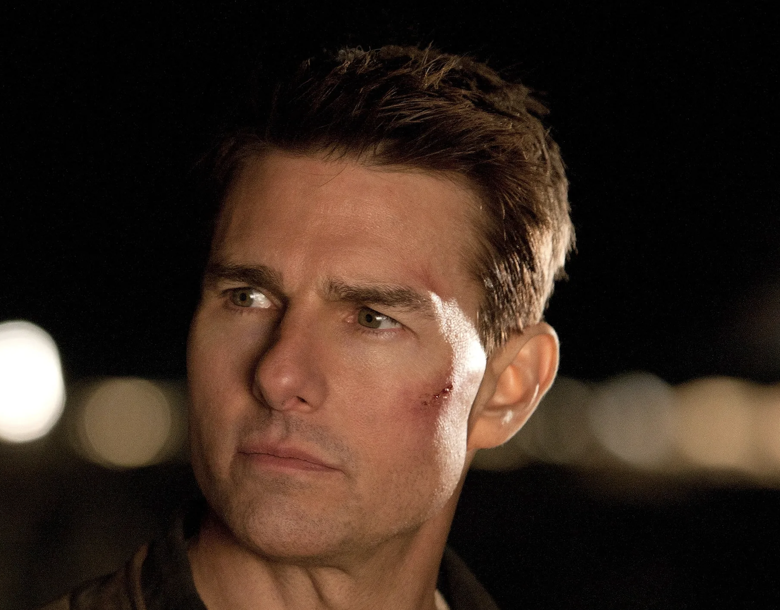 Tom Cruise with a slight bruise on his face, wearing a brown leather jacket, looking off to the side in a nighttime setting