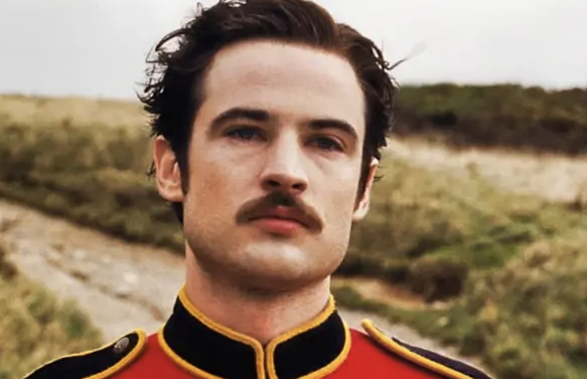 A person wearing a military-style jacket with a mustache stands outdoors with a serious expression. No identifiable celebrities are present