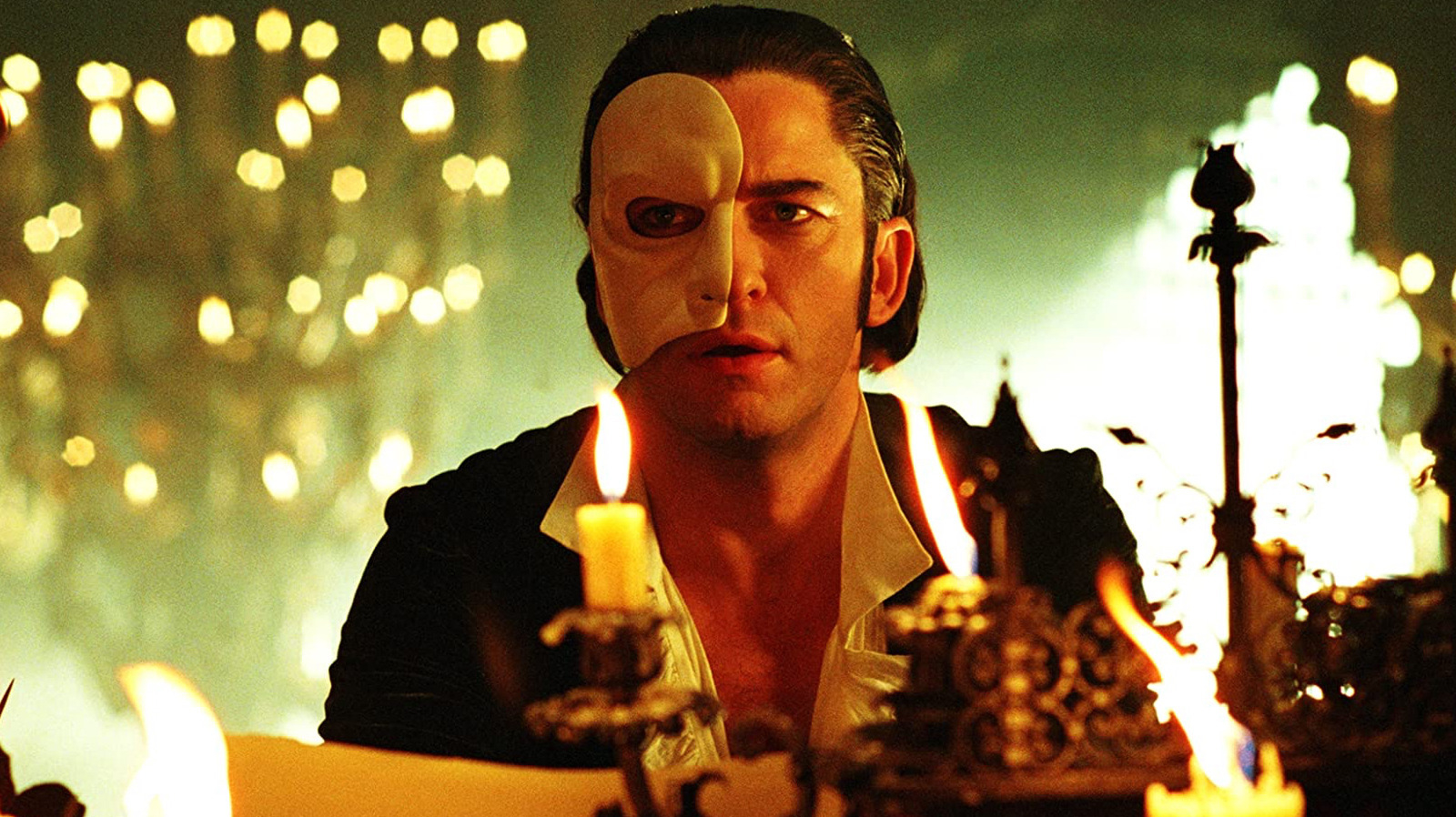 A scene from "The Phantom of the Opera" with Gerard Butler wearing half a white mask, positioned behind lit candles in a dimly lit setting