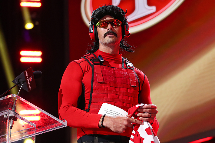 Dr. DisRespect, wearing a red tactical vest, sunglasses, and a wig, stands on stage holding a jersey and a speech