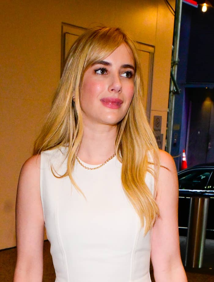 Emma Roberts is wearing a sleeveless, light-colored dress and a delicate necklace as she looks off to the side