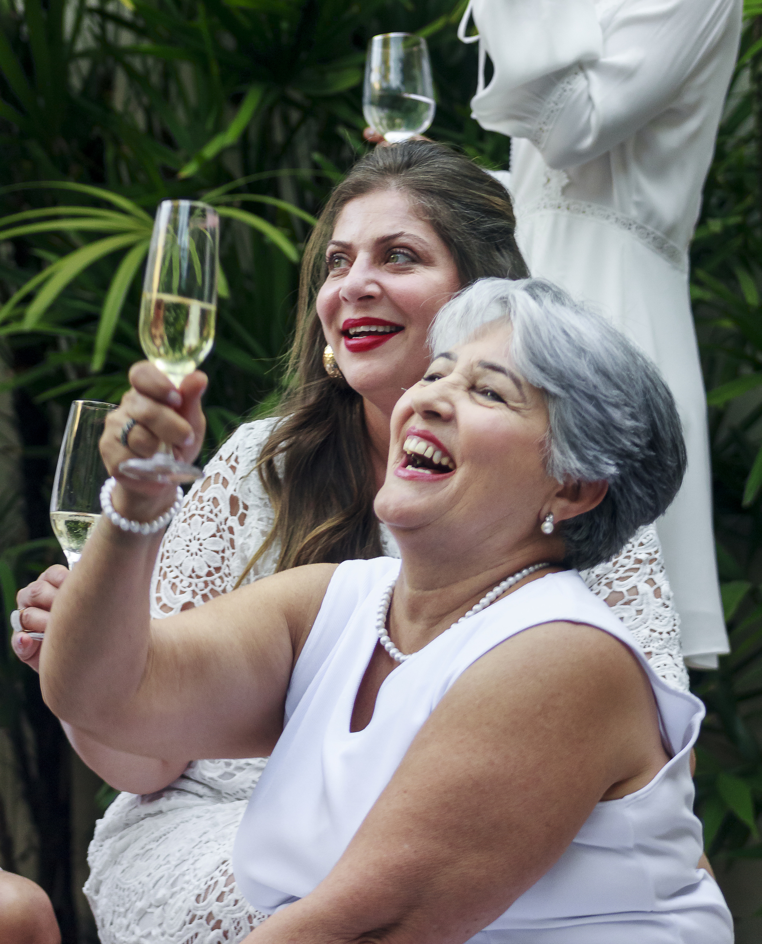 Two women in elegant white dresses, holding champagne flutes and laughing joyfully at a wedding