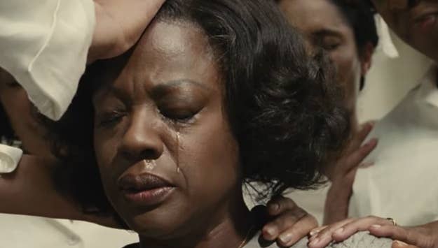 A woman, with tears on her face, is comforted by multiple hands placed gently on her head and shoulders