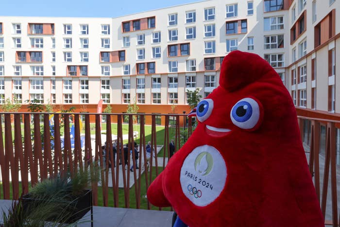 The image shows the Paris 2024 Olympic mascot standing on a balcony overlooking a courtyard of a modern building complex