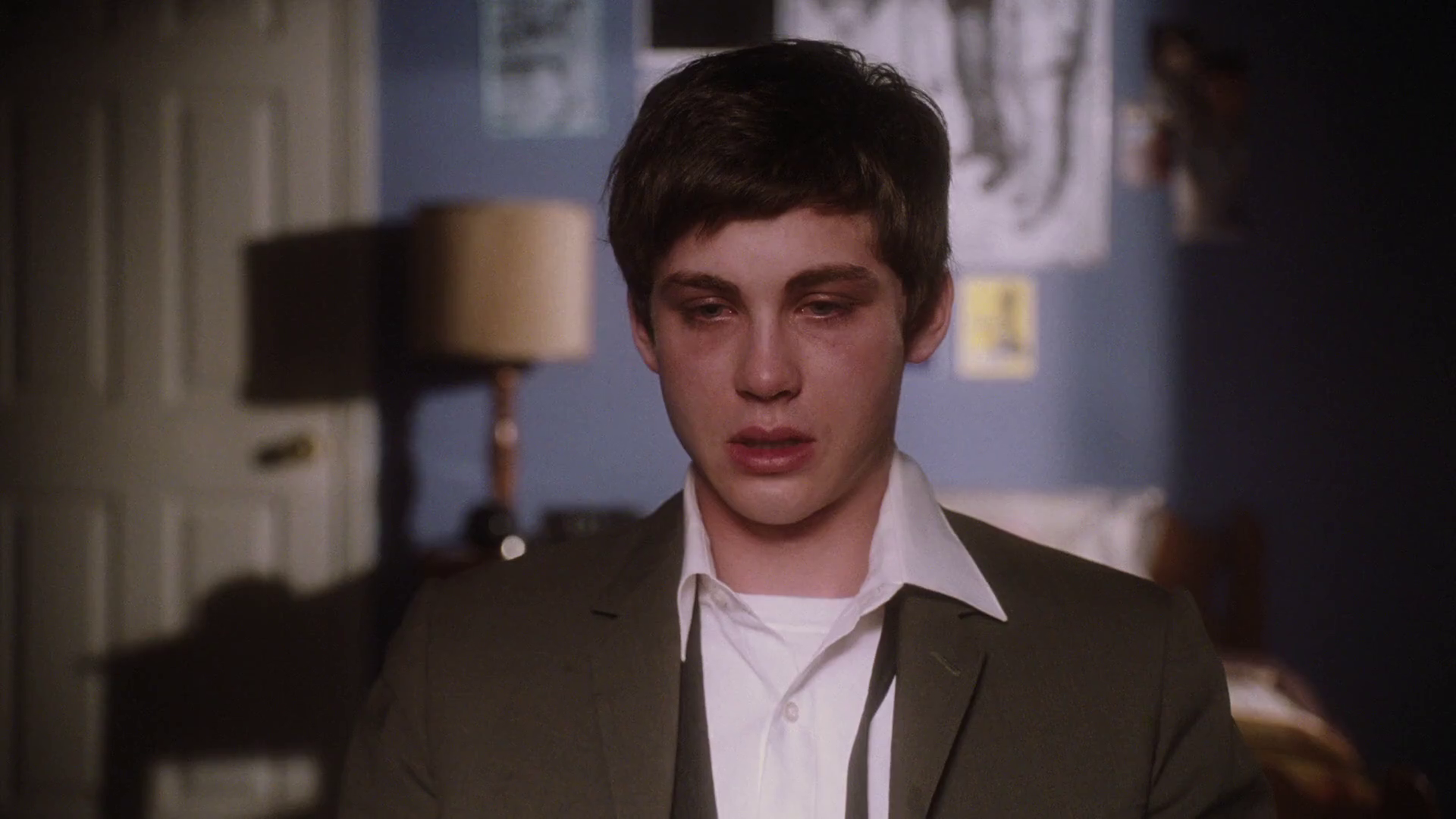 A person with short dark hair is wearing a white shirt and a suit jacket. They appear to be emotional or crying in a living room or bedroom setting