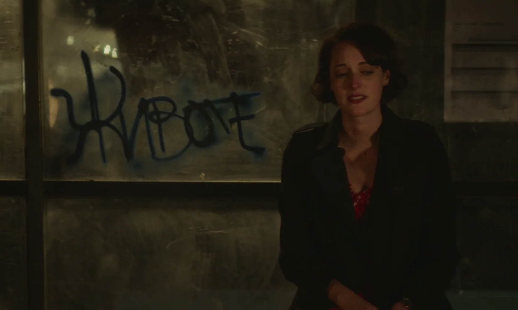 Phoebe Waller-Bridge sits against a glass wall with graffiti in the background. She wears a dark jacket over a red top