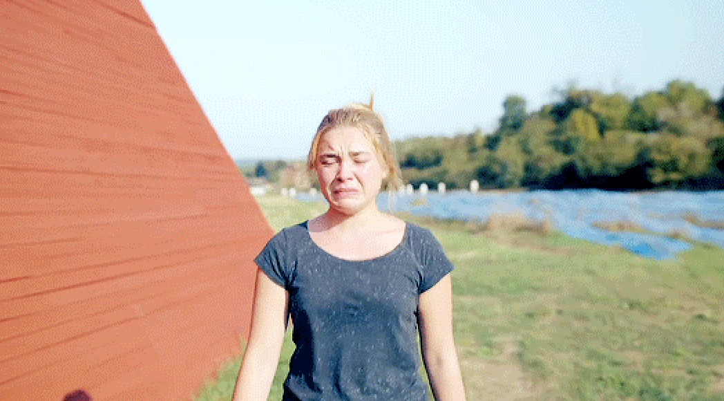Florence Pugh crying while standing outdoors next to a red structure with trees and fields in the background