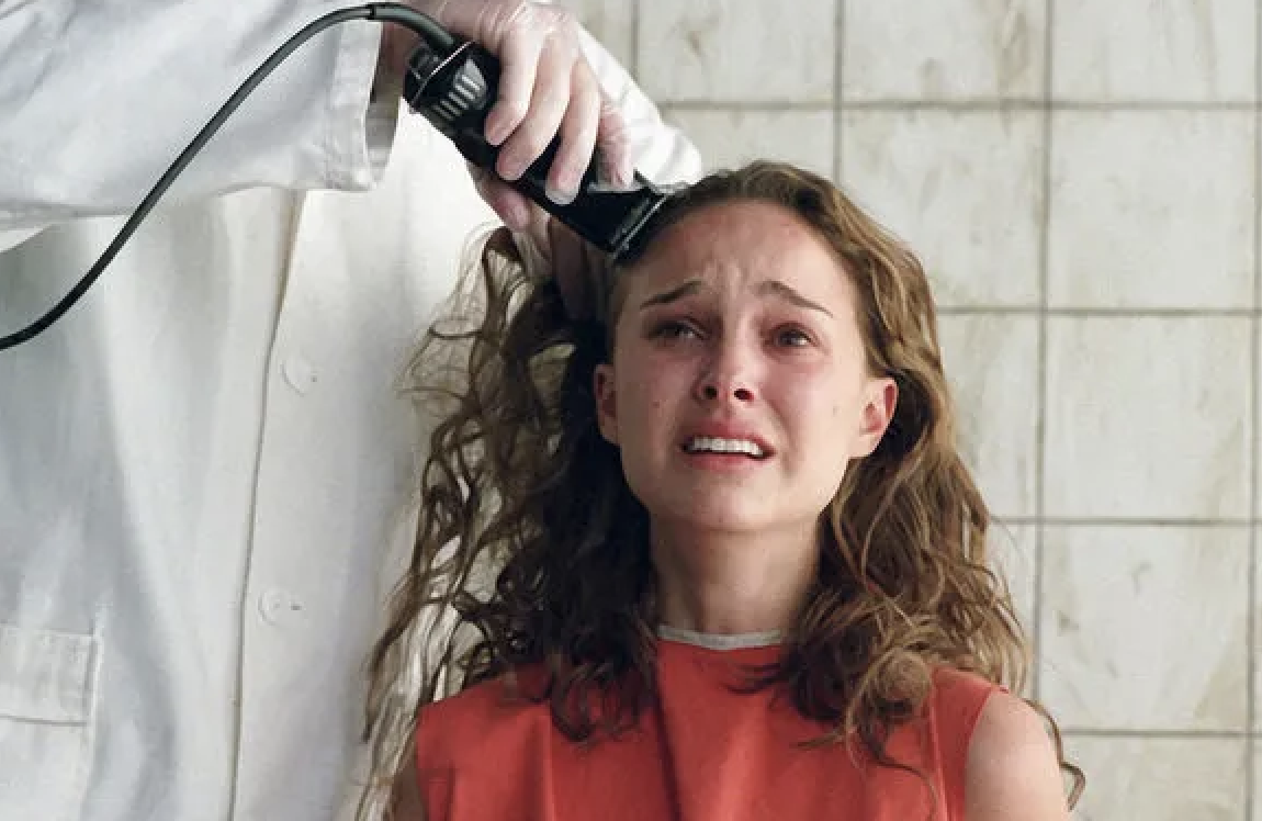 A person in a white coat shaves the head of Natalie Portman, who appears emotional, in a scene from a film
