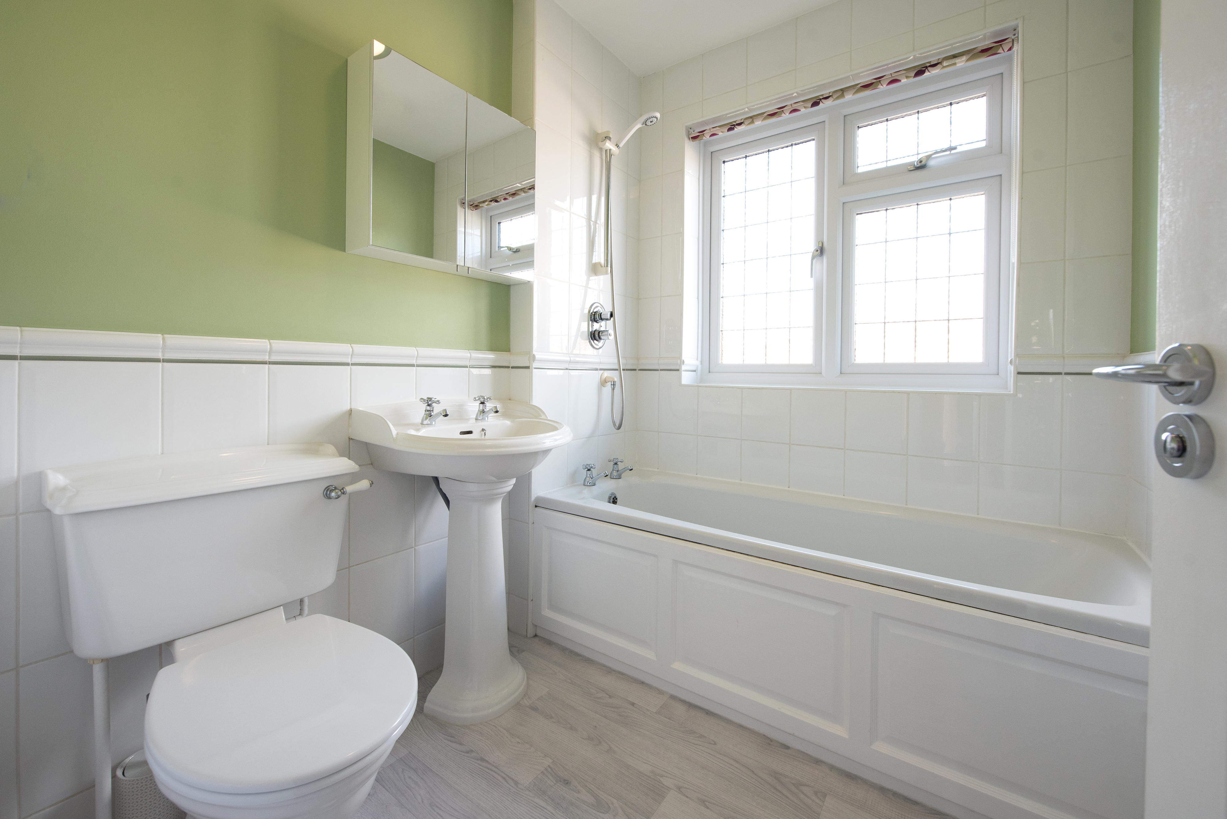 A clean and tidy bathroom with a pedestal sink, toilet, and bathtub next to a window. Light shines through the window, illuminating the space