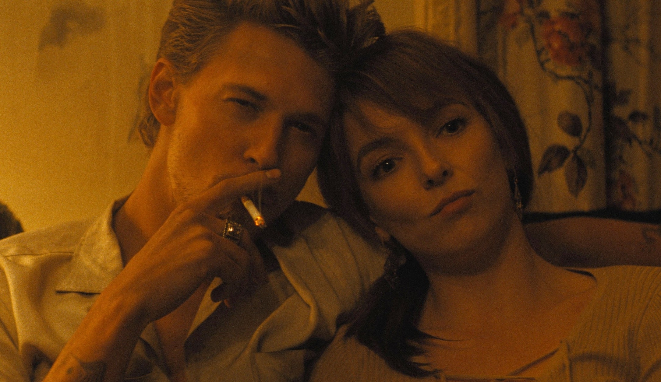 Austin Butler and Jodie Comer sit closely together on a couch; Austin smokes a cigarette while Jodie looks into the distance, both appearing contemplative
