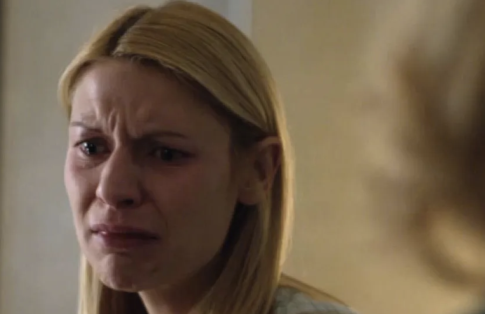 A woman with long blonde hair, wearing a serious expression, appears distressed in an indoor setting