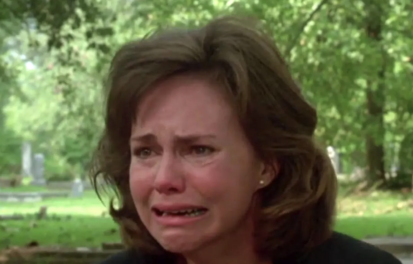 Sally Field is outdoors, crying with a distressed expression, possibly from an emotional scene in a movie. Green foliage is blurry in the background