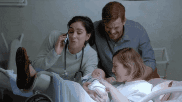 Sarah Silverman and Michael Gladis joyfully gather around a new mother holding her newborn in a hospital scene. Sarah talks on the phone