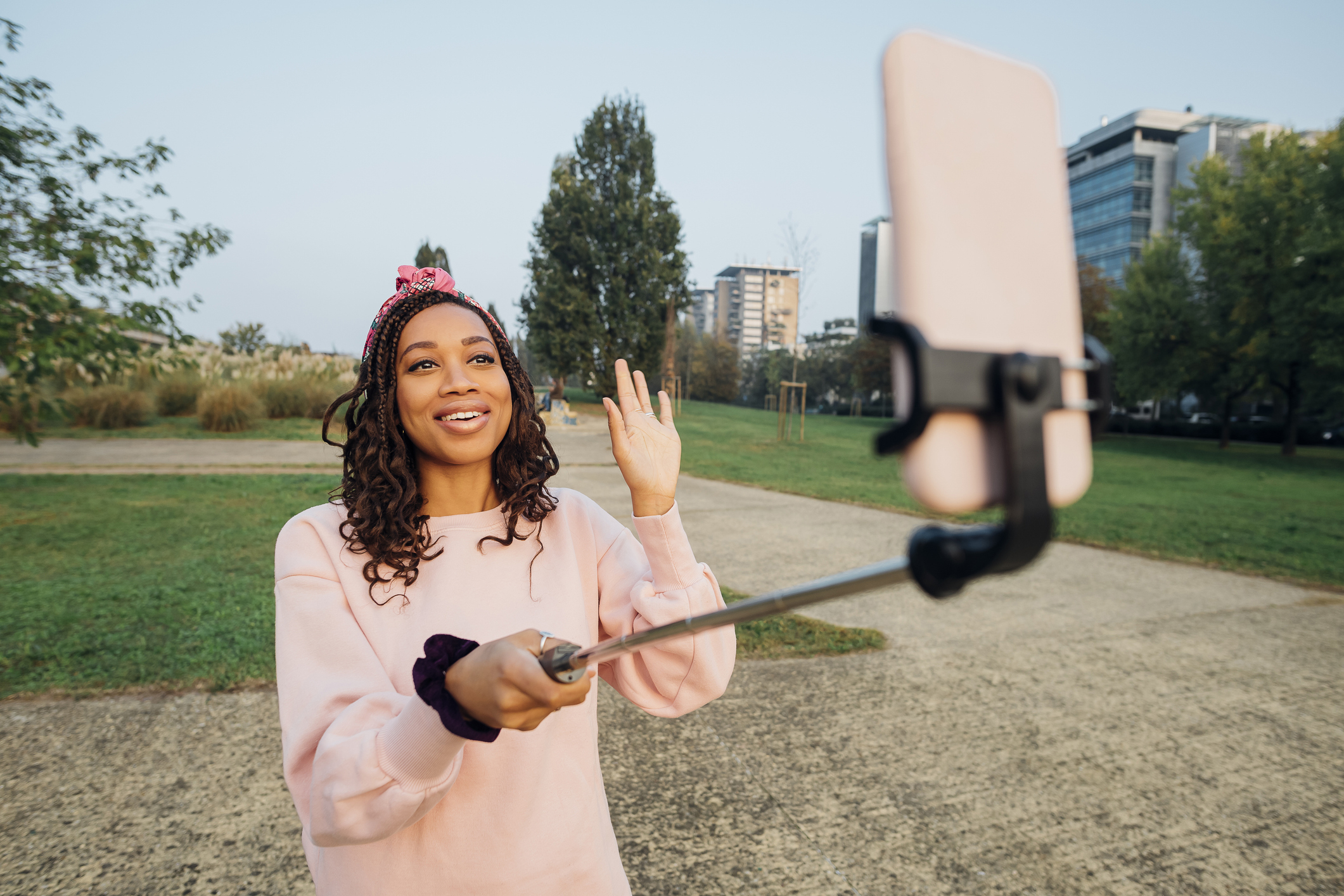 A woman stands outdoors in a park, smiling and waving while taking a selfie with a phone mounted on a selfie stick. She wears casual clothing and a headband
