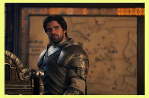 Criston Cole in medieval armor with a sword stands in front of a blurred map background