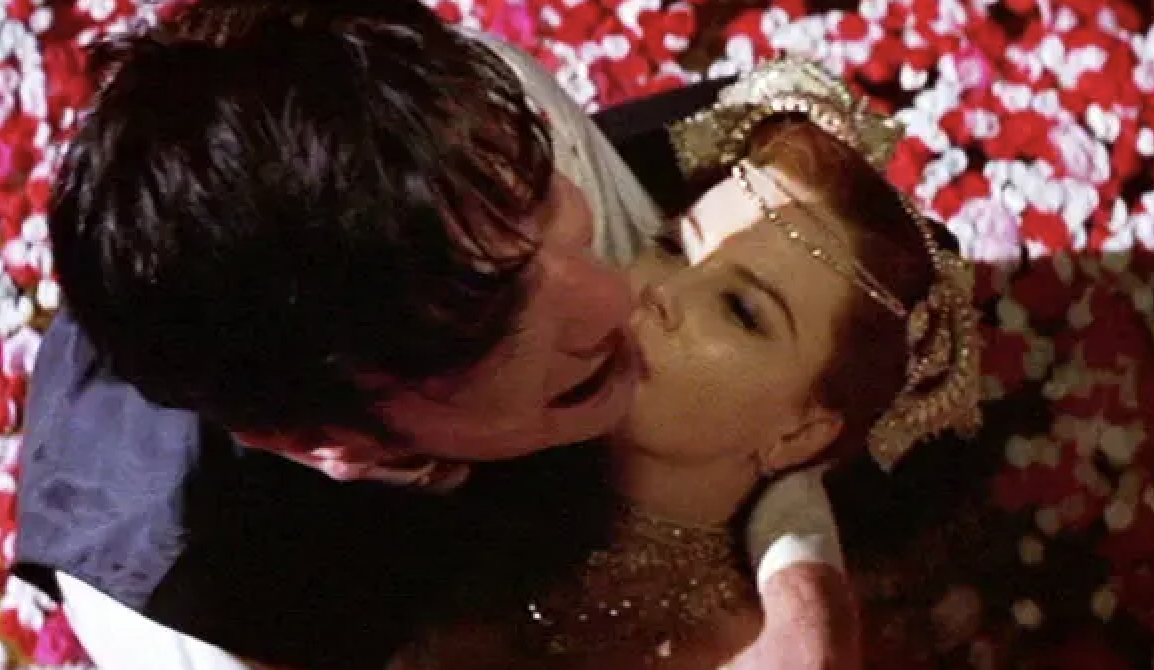 A couple embraces and kisses against a backdrop of rose petals. The woman is wearing an ornate headpiece and jewelry. Both appear intense