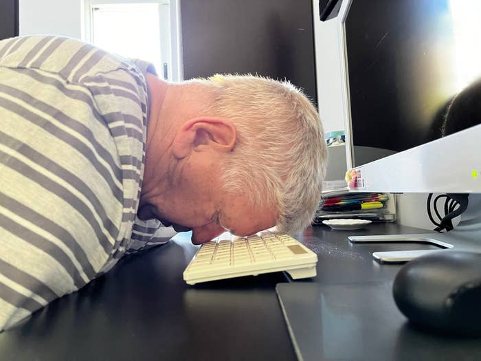 Older man with short hair rests his head on a keyboard at a desk in front of a computer screen