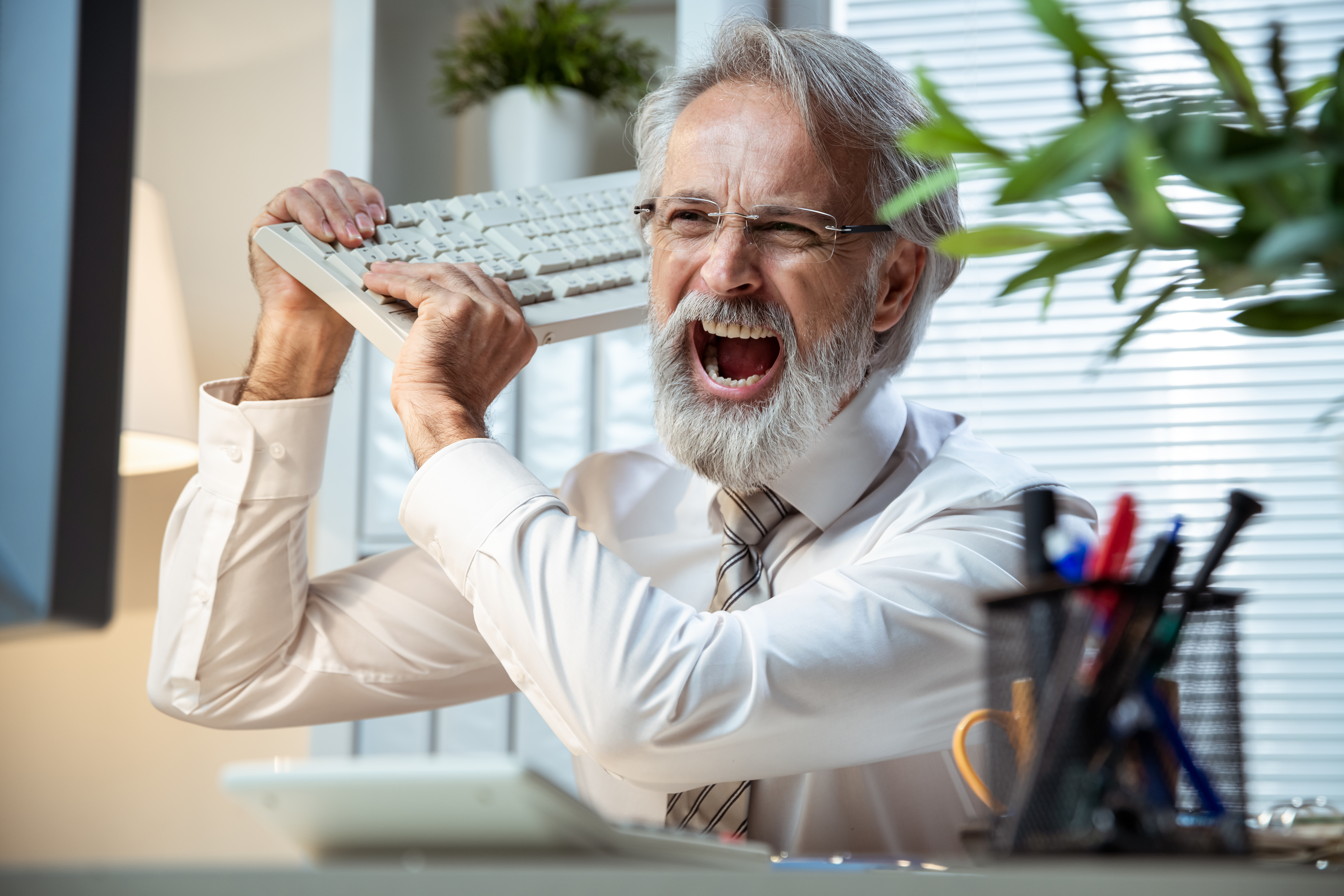 A man in an office, wearing a shirt and tie, angrily holding a keyboard above his head, about to smash it