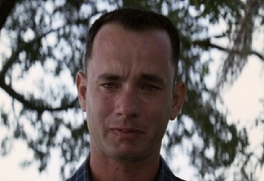 Man with short hair and a sad expression stands outdoors with trees in the background