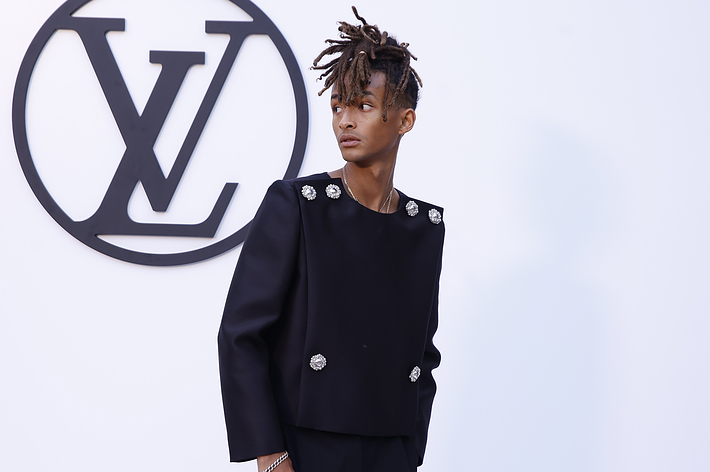 Jaden Smith on a red carpet, wearing a stylish black suit with embellishments, standing in front of a large Louis Vuitton logo