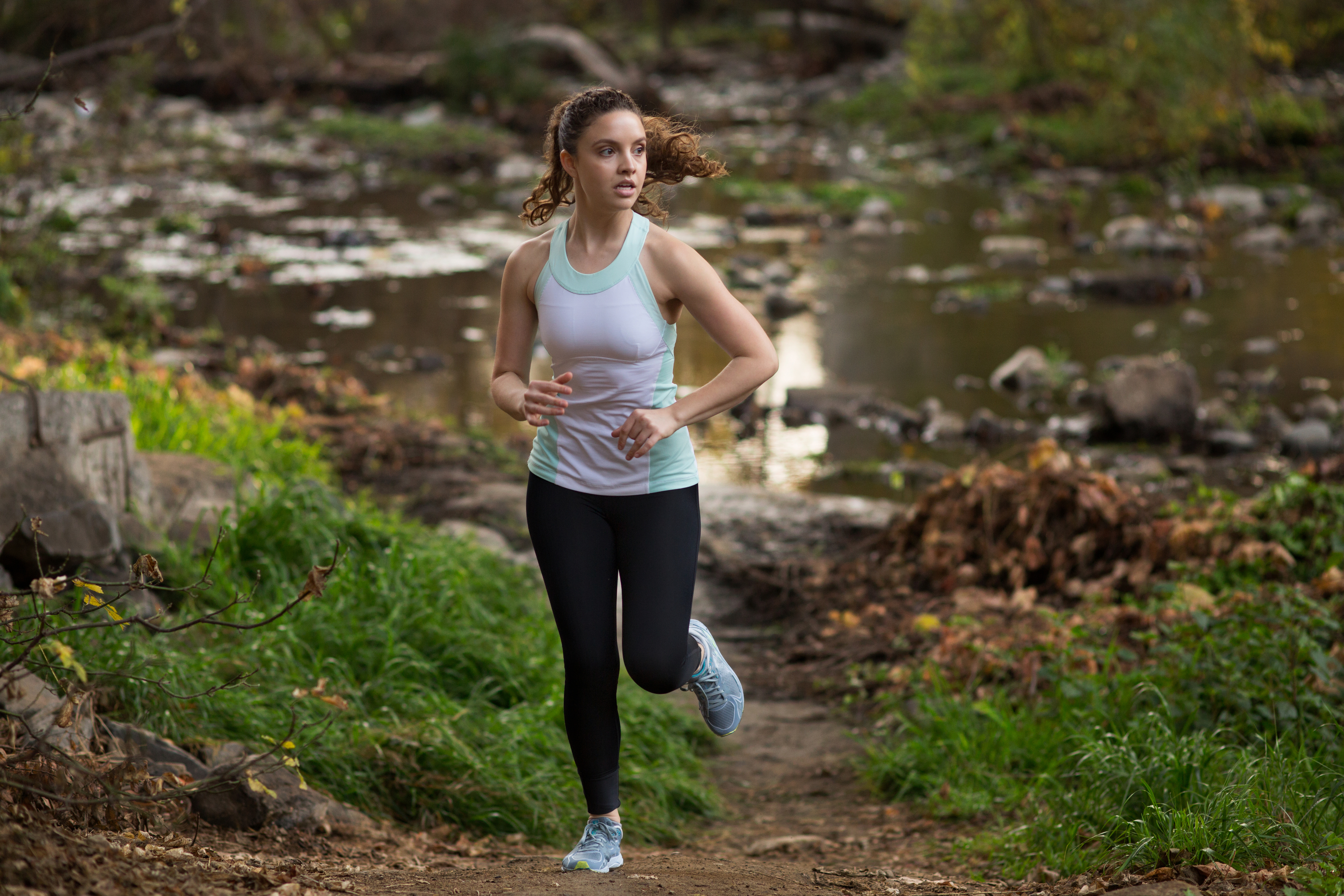 A woman with curly hair wearing a sleeveless athletic top and leggings is running on a trail in a forested area near a stream
