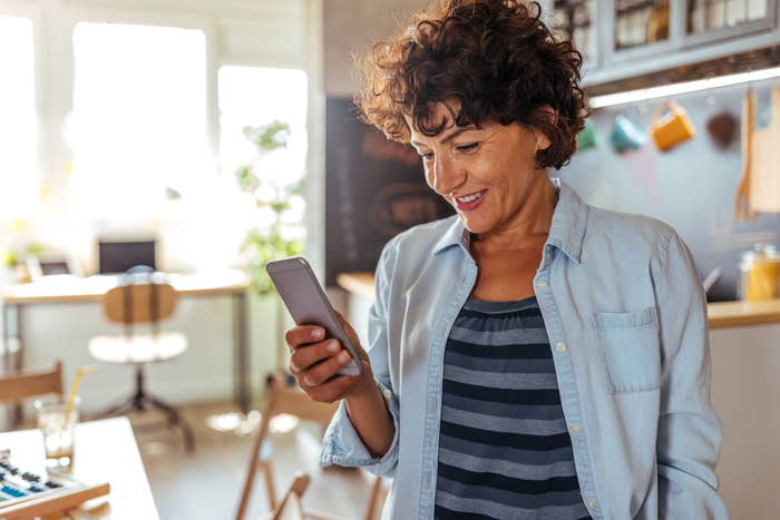 A person with curly hair smiles while looking at a smartphone in a brightly lit kitchen