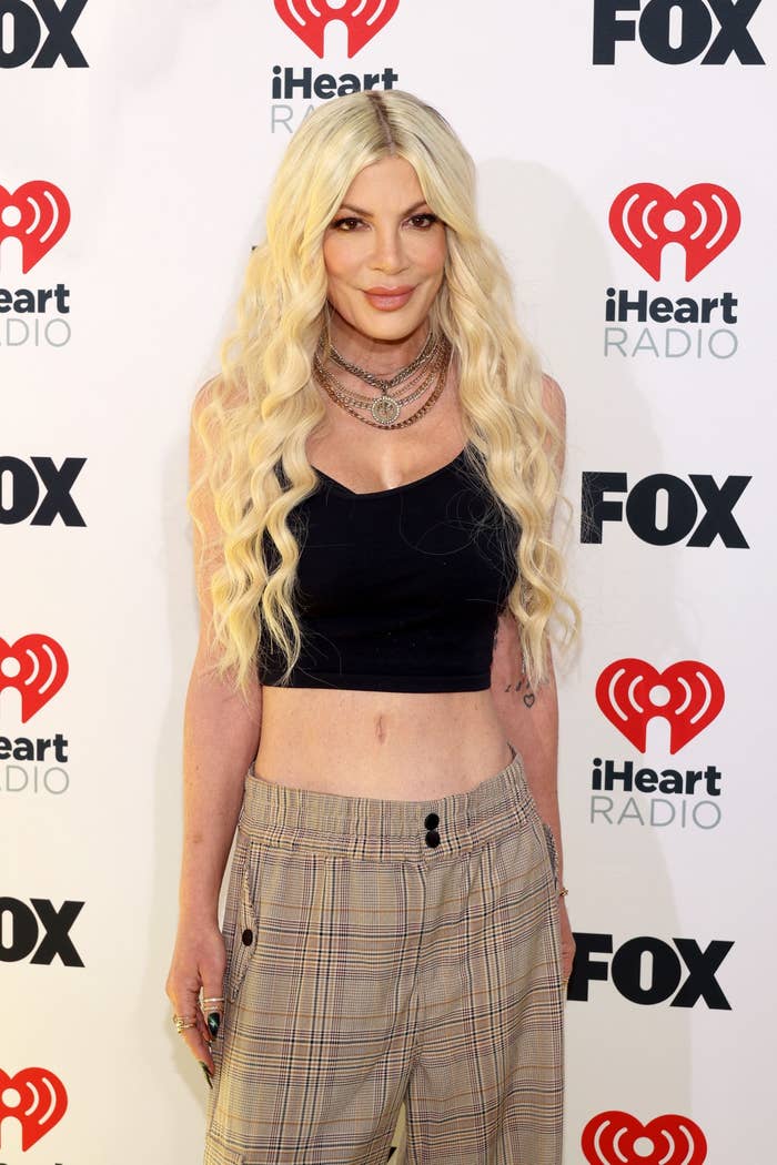 Tori Spelling on the red carpet at an iHeartRadio Fox event, wearing a crop top and plaid pants, with long wavy hair and layered necklaces