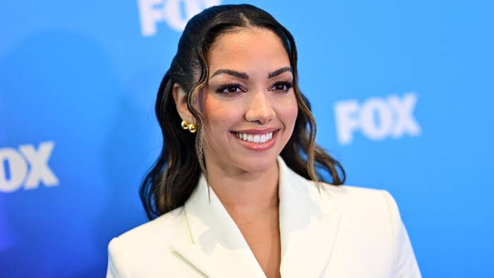 A woman with long wavy hair smiles at a FOX event, wearing a stylish white blazer with gold earrings