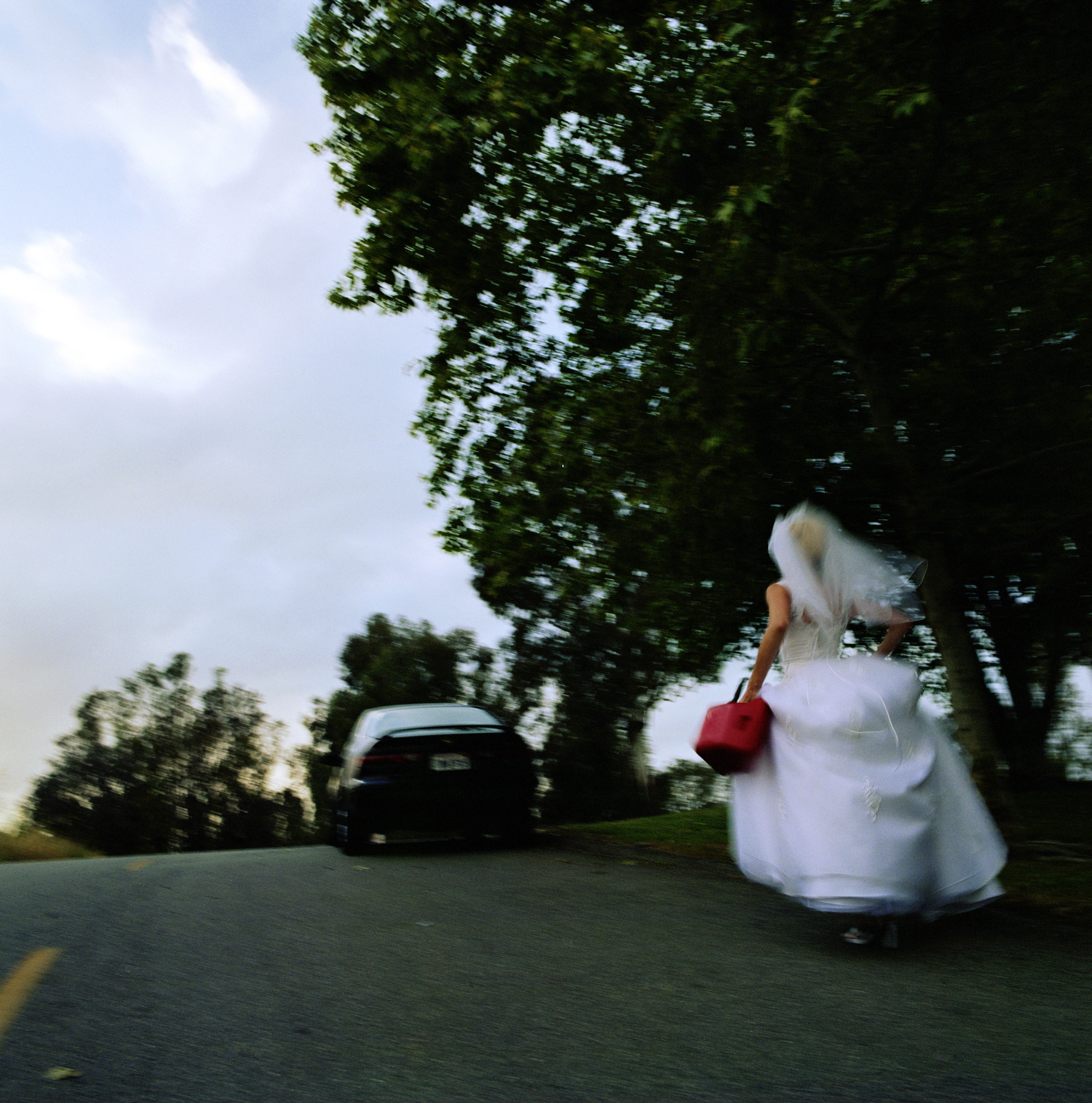 A person in a wedding dress holding a red bag is running down a road with trees and a parked black car in the background
