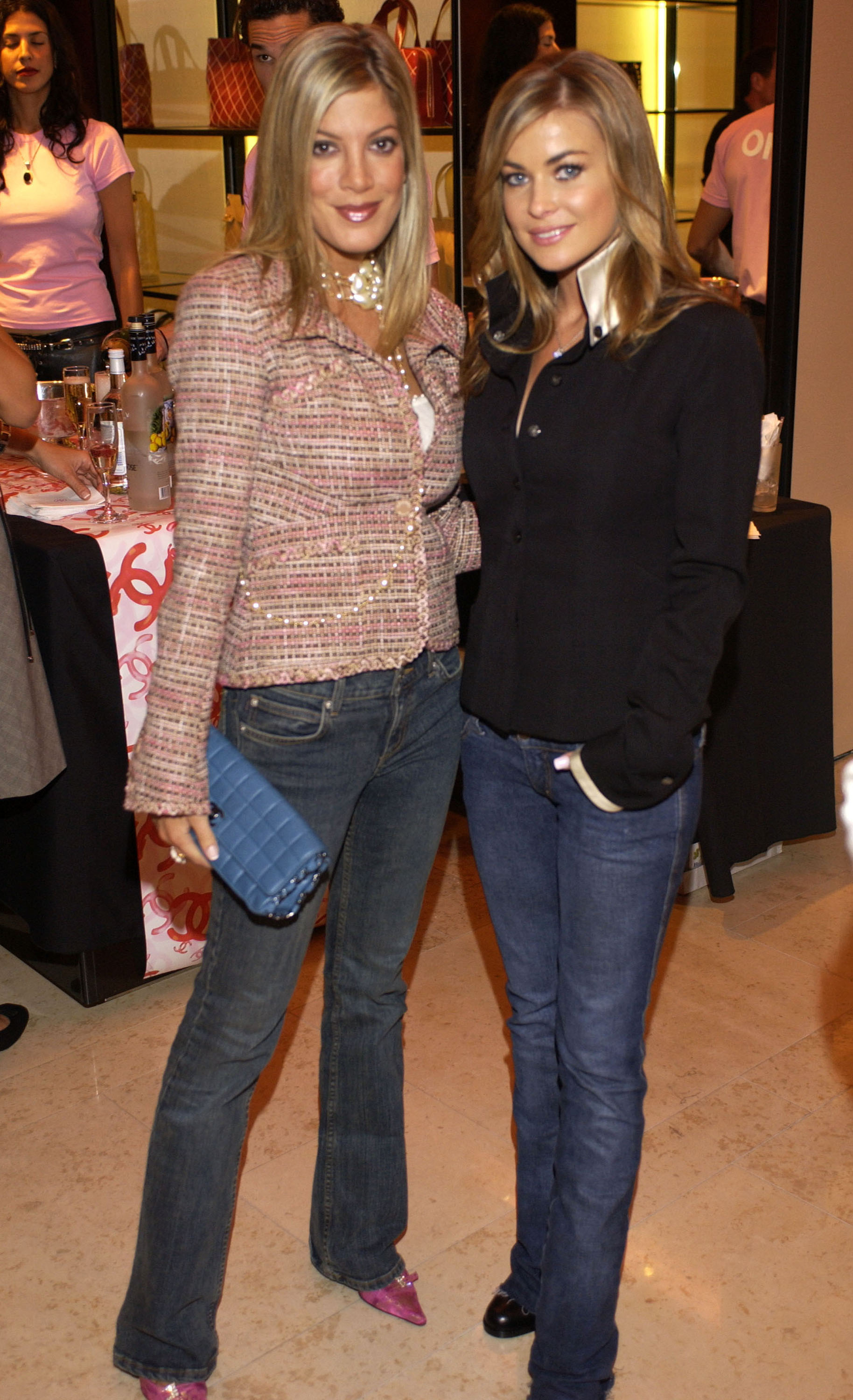 Tori Spelling and Carmen Electra posing together at an event, both wearing jeans and jackets. Tori holds a clutch