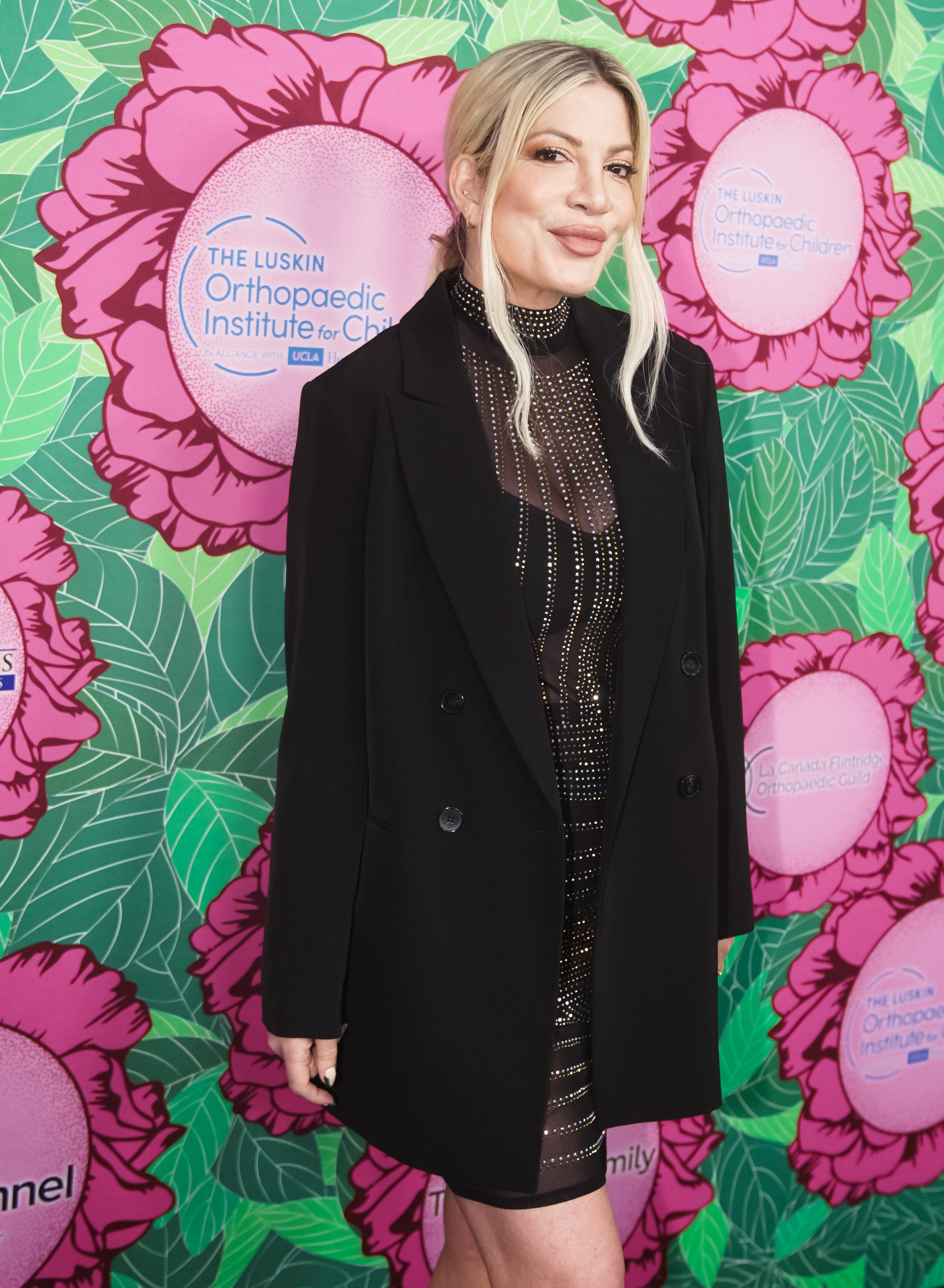Tori Spelling in a dark blazer over a sparkling, sheer dress at an event with a floral backdrop