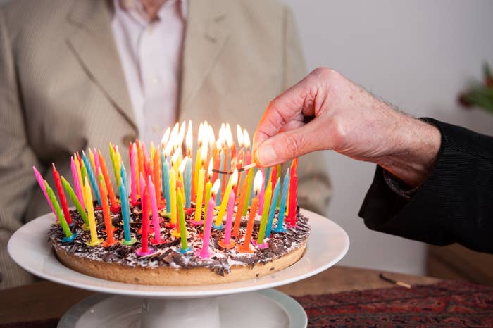 A person lights numerous colorful candles on a birthday cake while another person stands in the background in a suit