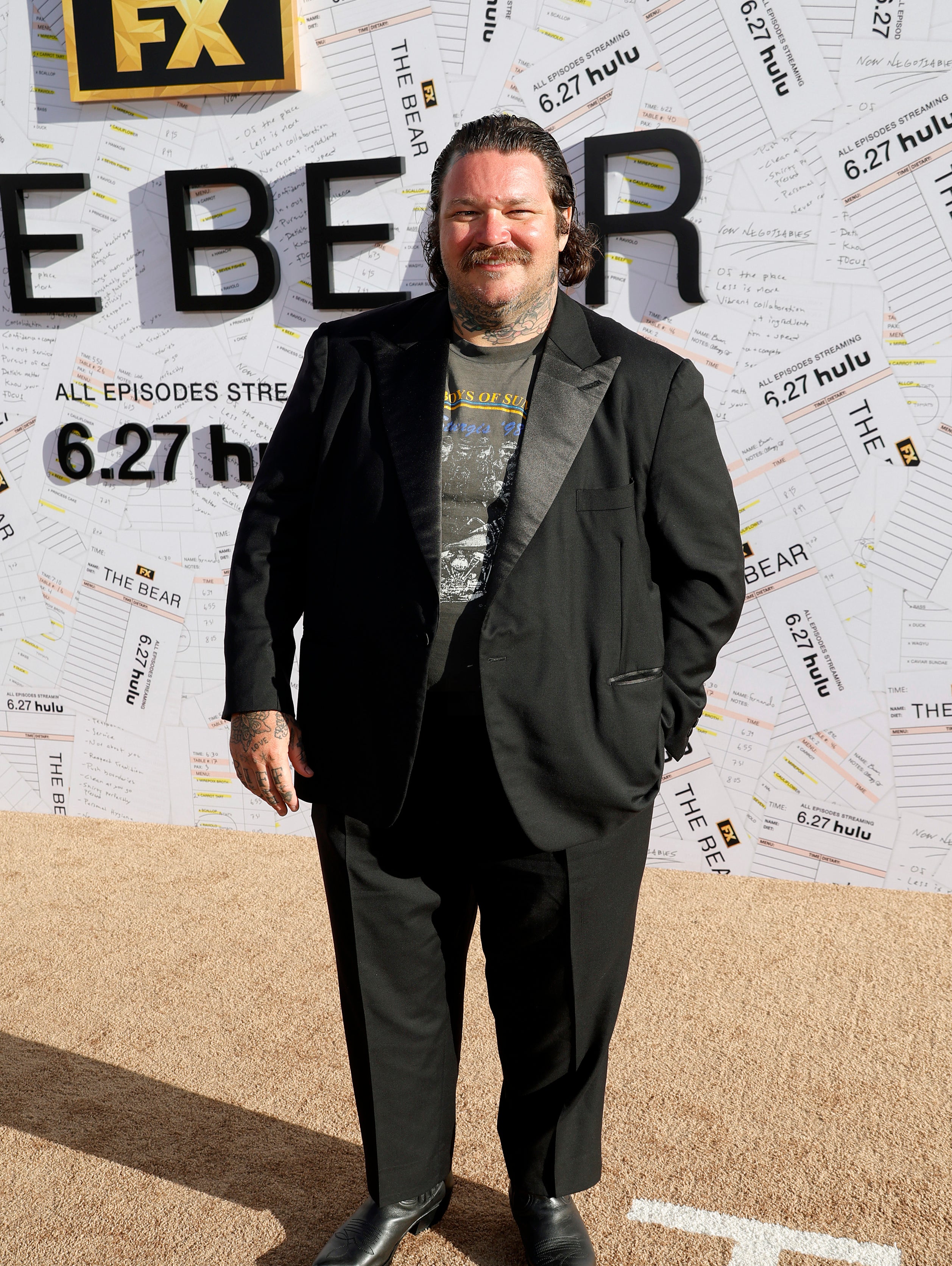 Matty Matheson at the FX show &quot;The Bear&quot; premiere, wearing a black blazer, graphic tee, black pants, and black shoes, standing in front of a promotional background