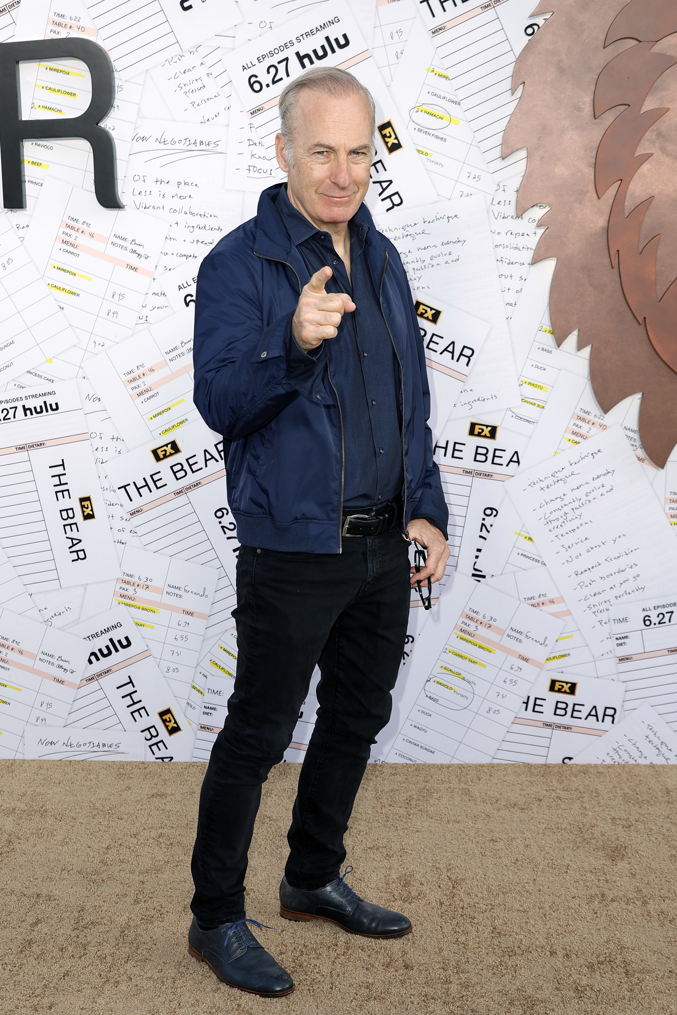 Bob Odenkirk points at the camera while standing in front of a backdrop featuring promotional material for Hulu's &quot;The Bear.&quot;