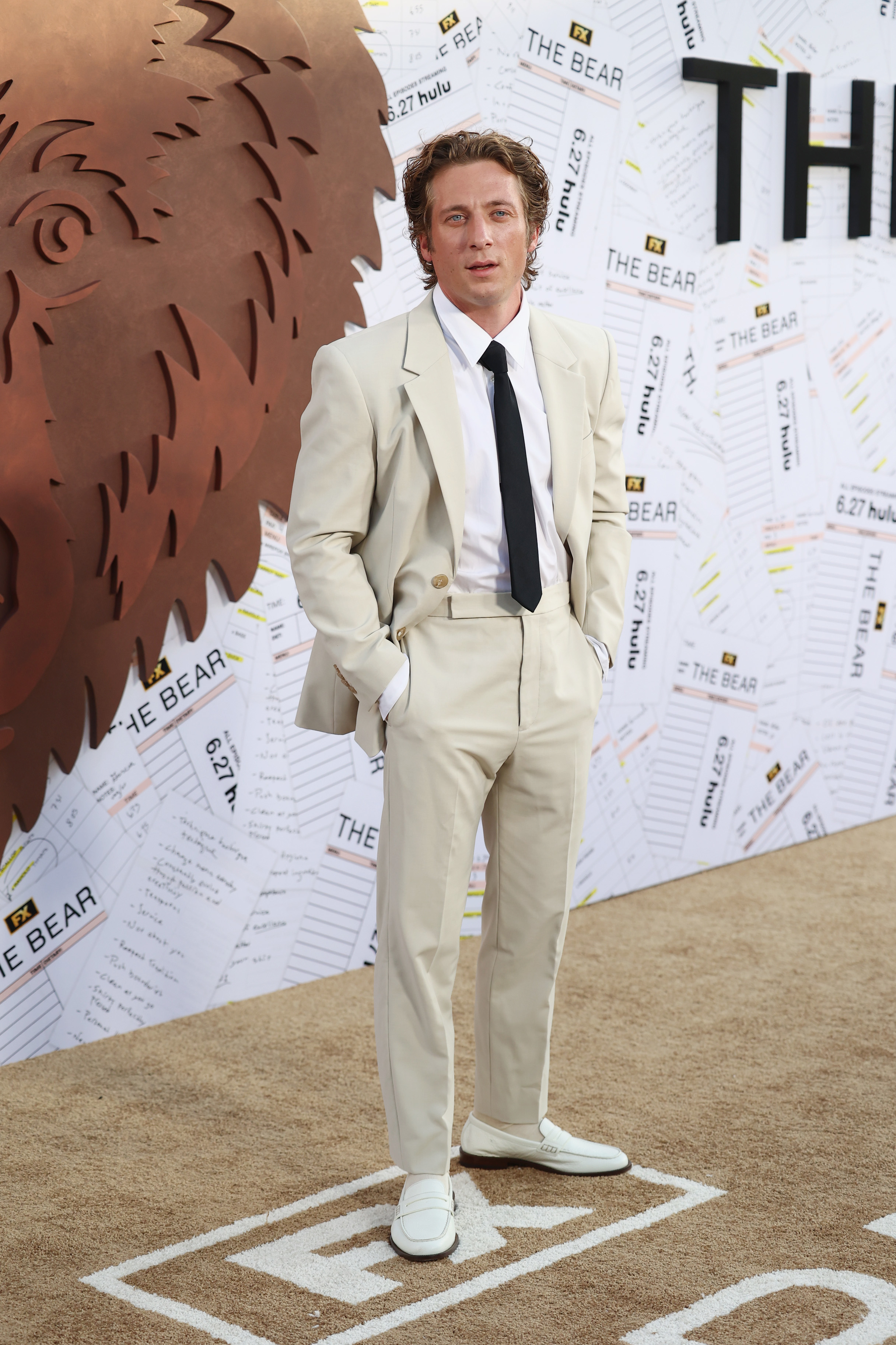 Jeremy Allen White at a promotional event for &quot;The Bear,&quot; wearing a white suit with a black tie and white loafers, standing against a backdrop of the show's theme