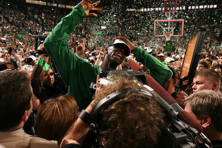 Basketball player celebrates victory on the court surrounded by fans and media, with confetti falling in the background