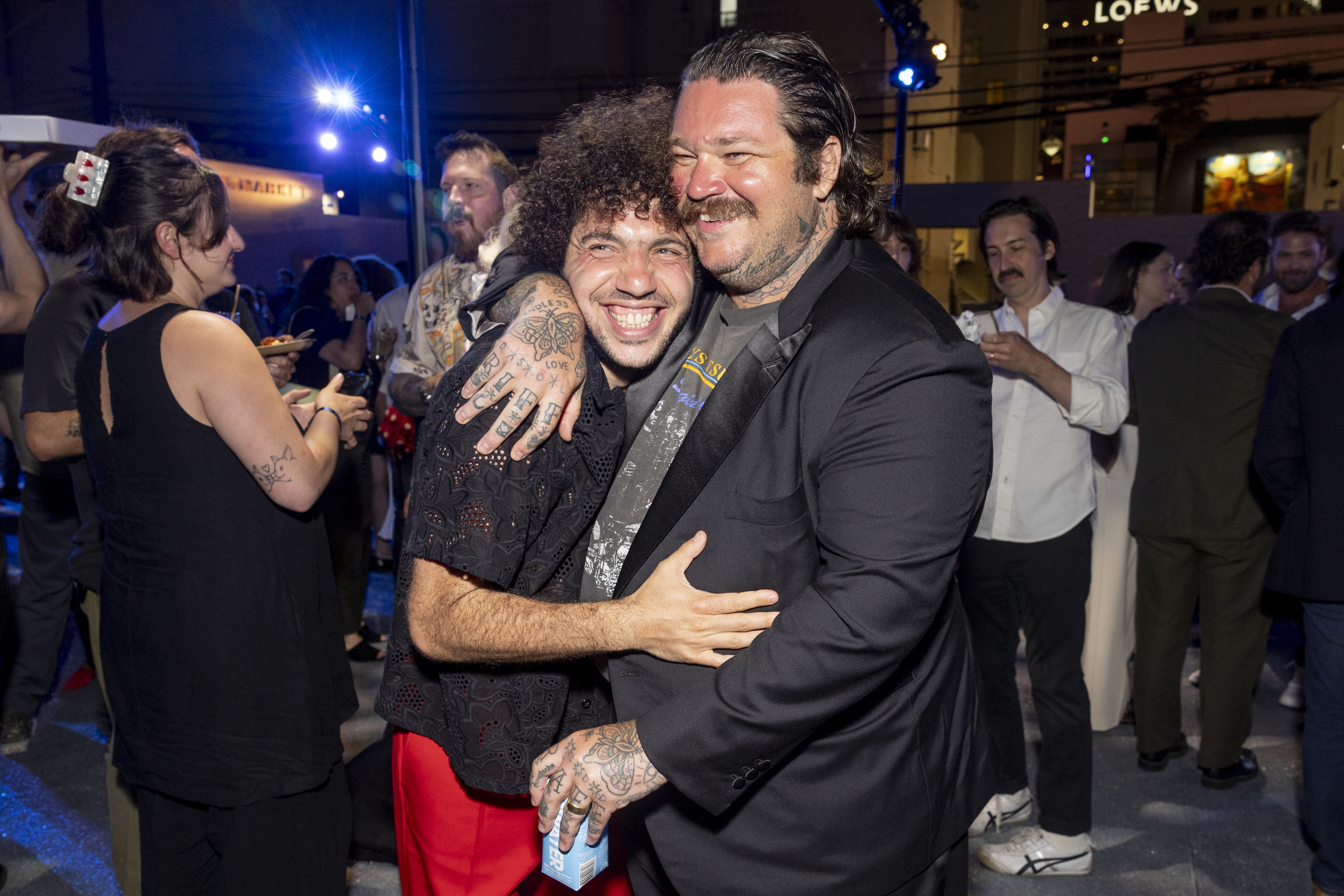 Benny Blanco and Matty Matheson hugging and smiling at a lively event, surrounded by other guests in the background. Benny wears a black shirt and red pants